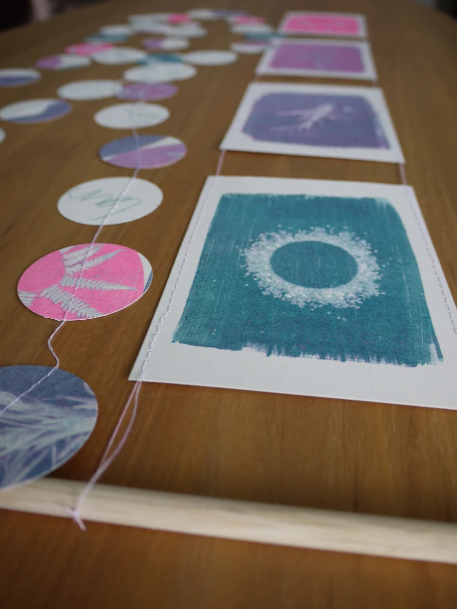 Everyday Enchantment - A close-up of a wooden table with a handmade cyanotype garland made of stitched circular paper cutouts,  alongside a riso print wall hanging on a wooden dowel.
