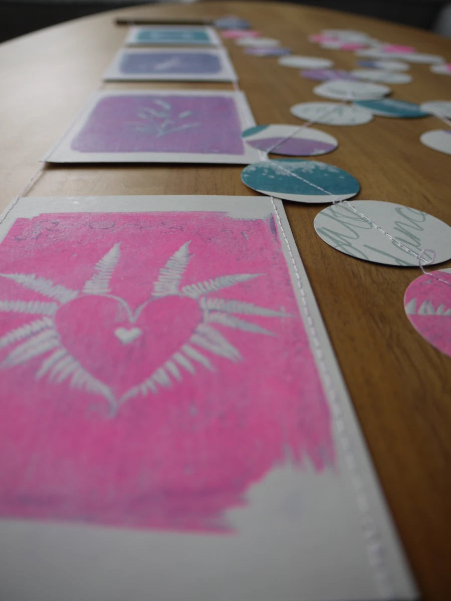 Everyday Enchantment - A close-up of a handmade riso print wall hanging beside a cyanotype garland. Riso printed with pink, purple, and teal designs, including a heart motif on a white card in the foreground.