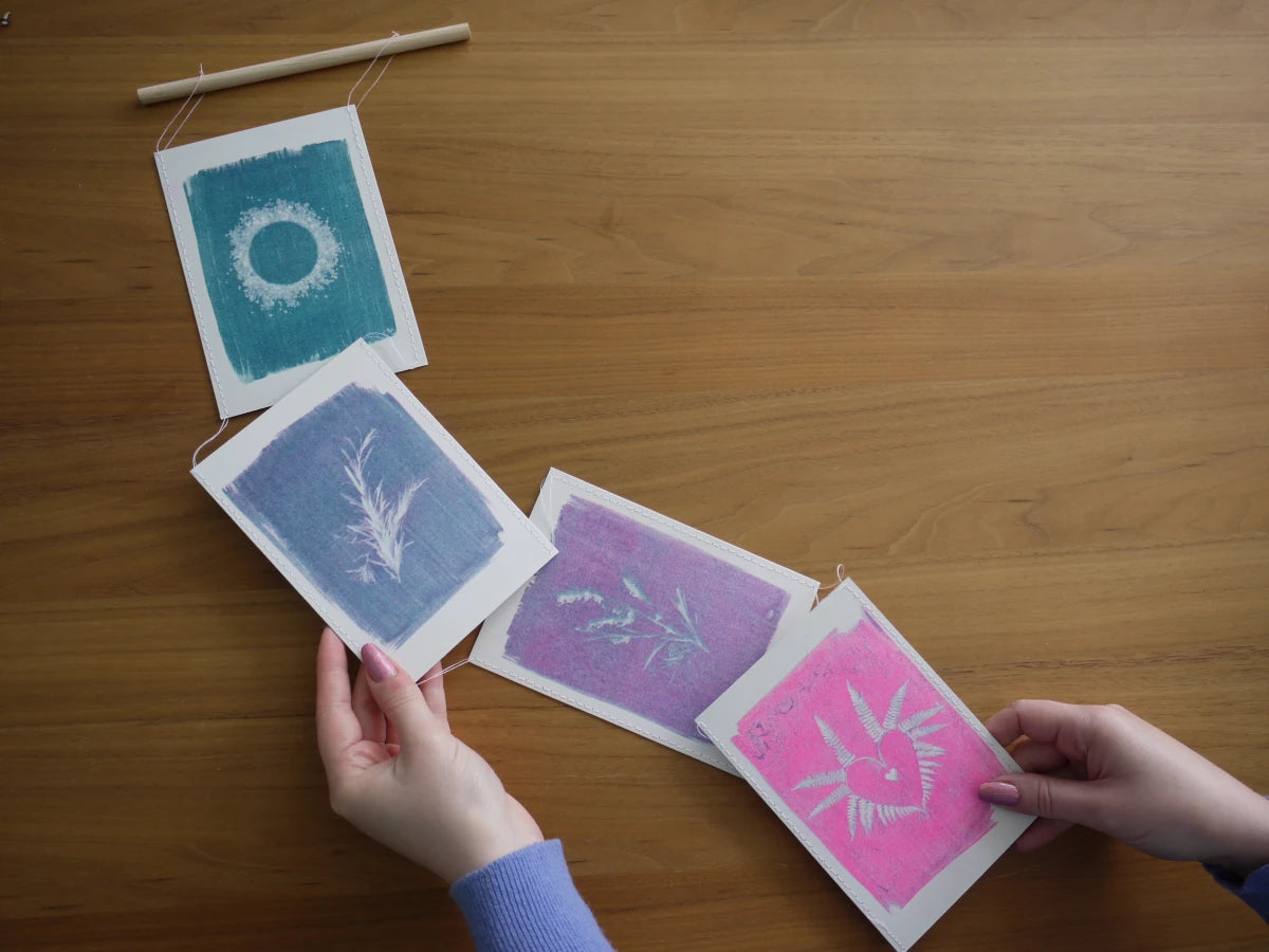 Everyday Enchantment - Four handmade cyanotype riso art prints in teal, blue, and pink are being held by hands over a wooden table, with a wooden dowel and thread visible at the top.
