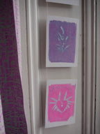 Everyday Enchantment - riso print wall hanging. Two small art prints hanging vertically on a white door. The top print is lavender/purple with a white plant design, and the bottom print is bright pink with a white sun-and-heart design.