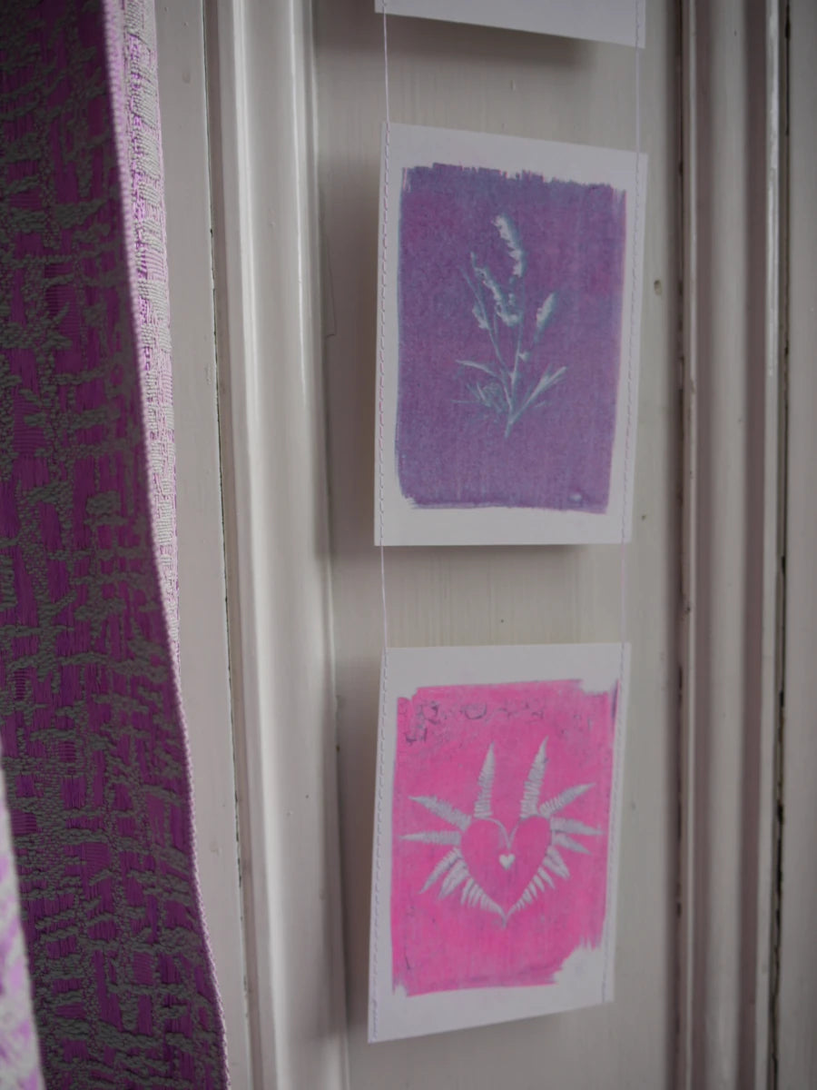 Everyday Enchantment - riso print wall hanging. Two small art prints hanging vertically on a white door. The top print is lavender/purple with a white plant design, and the bottom print is bright pink with a white sun-and-heart design.