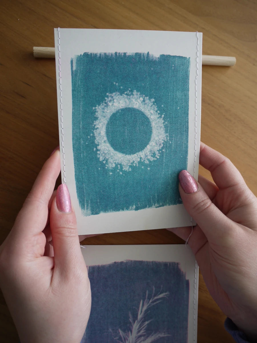 Everyday Enchantment - A person holds a small unique cyanotype as part of a riso print wall hanging stitched onto a wooden dowel. Art print featuring a white ring design on a teal-blue background and another print partially visible below on a wooden table.