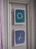 Everyday Enchantment - riso print wall hanging from original unique cyanotypes. Two prints hanging on a white door from a wooden dowel. The top print features a teal blue rectangle with a white circular motif, and the bottom print features a botanical rosemary sprig design.