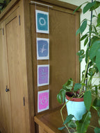 Everyday Enchantment - A wooden cabinet in a home setting with four art prints in blue, purple, and pink to make a riso print wall hanging next to a potted green plant.