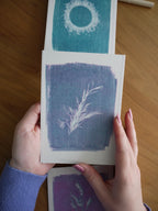 Everyday Enchantment - A person's hands holding a cyanotype riso print as part of a riso print wall hanging stitched onto a wooden dowel. Art print with a white rosemary sprig design on a textured blue background, with other art prints visible on a wooden table.