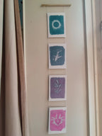 Everyday Enchantment - Four vertical unique cyanotypes riso printed and stitched together to create a riso print wall hanging. Botanical art prints in teal, blue-green, purple, and pink, hanging from a wooden dowel on a beige wall next to a curtain.