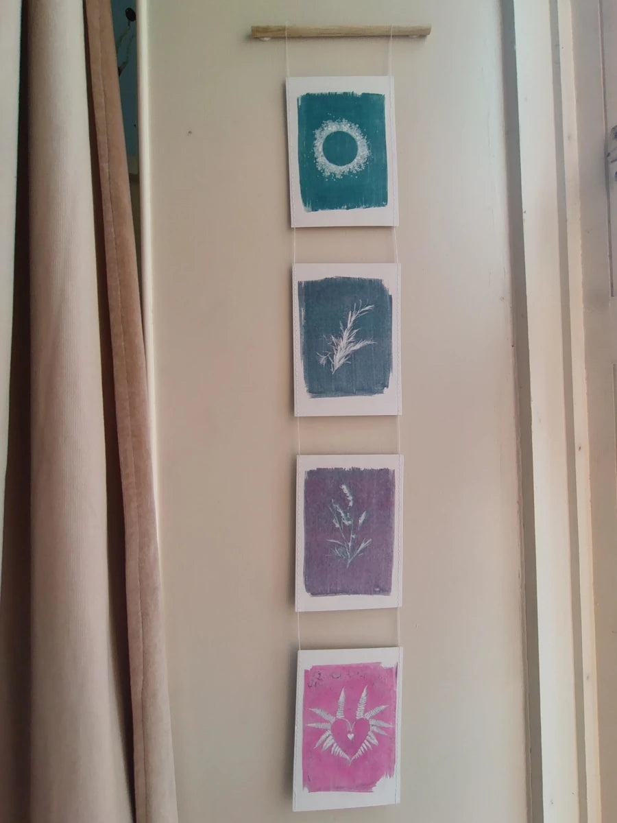 Everyday Enchantment - Four vertical unique cyanotypes riso printed and stitched together to create a riso print wall hanging. Botanical art prints in teal, blue-green, purple, and pink, hanging from a wooden dowel on a beige wall next to a curtain.