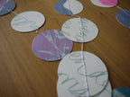 A close-up of a circular garland made of white paper circles with riso print pastel designs, stitched together with white thread and sat along a wooden tabletop.