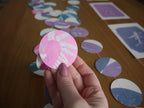 A hand with glittery pink nails holds a circular pink and white fern-patterned circle, with with the rest of the riso print garland scattered on a wooden surface in the background.