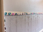 A decorative border of riso print cyanotype paper circle garland in teal, pink, white, and blue attached to a dado rail above a gey embossed floral wallpaper panel in an indoor room.