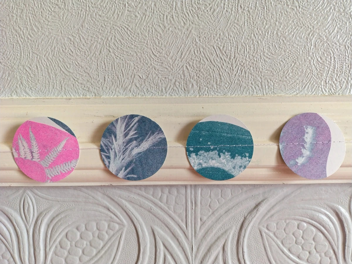Four circular riso print cyanotype shapes with different botanical and abstract patterns in pink, blue, teal, and purple, stitched together and hung in a hallway.