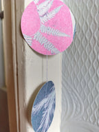 A decorative hanging garland with two circular shapes riso printed with white fern and botanical patterns, shown hanging against a textured white wall.