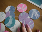 A close-up of a handmade circular garland made of stitched riso print  circles with pastel botanical prints in pink, purple, blue, and white, arranged on a wooden table with a person's hand visible.