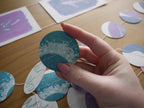 A hand with pink nail polish holds a round teal and white cyanotype riso print paper garland circle on a wooden table, with the rest of the garland on the table.