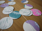 Circular paper garland with various riso printed cyanotype designs in pink, teal, purple, and white, stitched together on a wooden surface.
