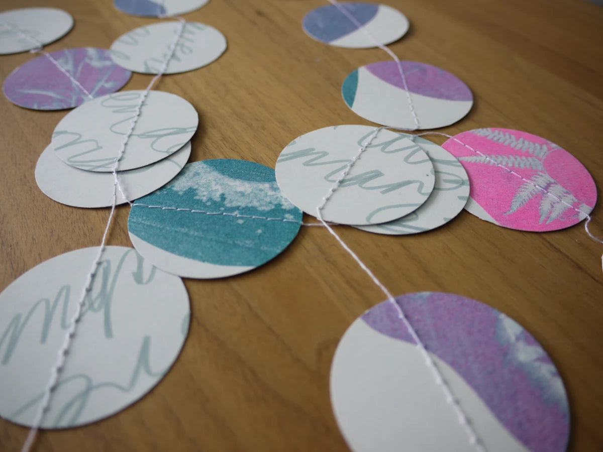 Circular paper garland with various riso printed cyanotype designs in pink, teal, purple, and white, stitched together on a wooden surface.
