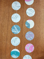 Circular paper riso print garland with abstract blue, teal, pink, and lavender cyanotype patterns hanging on a wooden surface.
