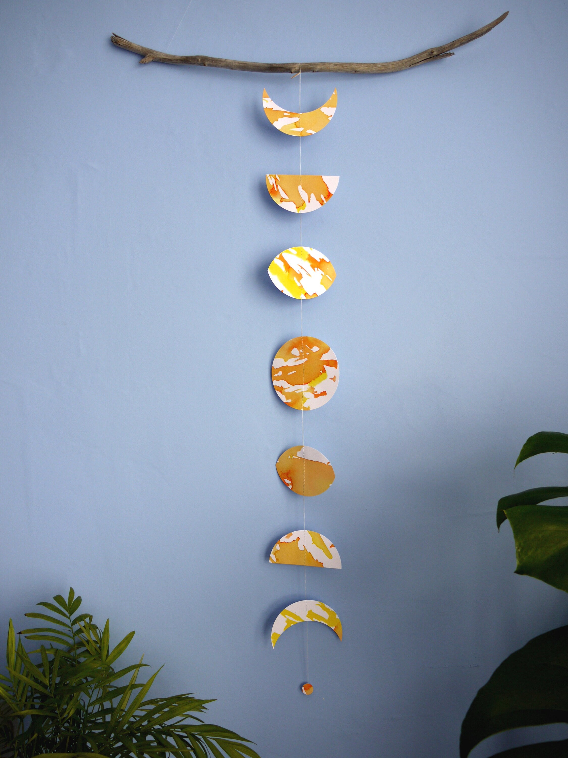 Flower Moon phases wall hanging. Eco friendly decor hand-painted with yellow, orange and gold ink. Decorative paper garland hanging from driftwood as bedroom wall decor, surrounded by houseplants