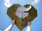 A giant gold origami heart held up with a blue sky and white clouds as the background