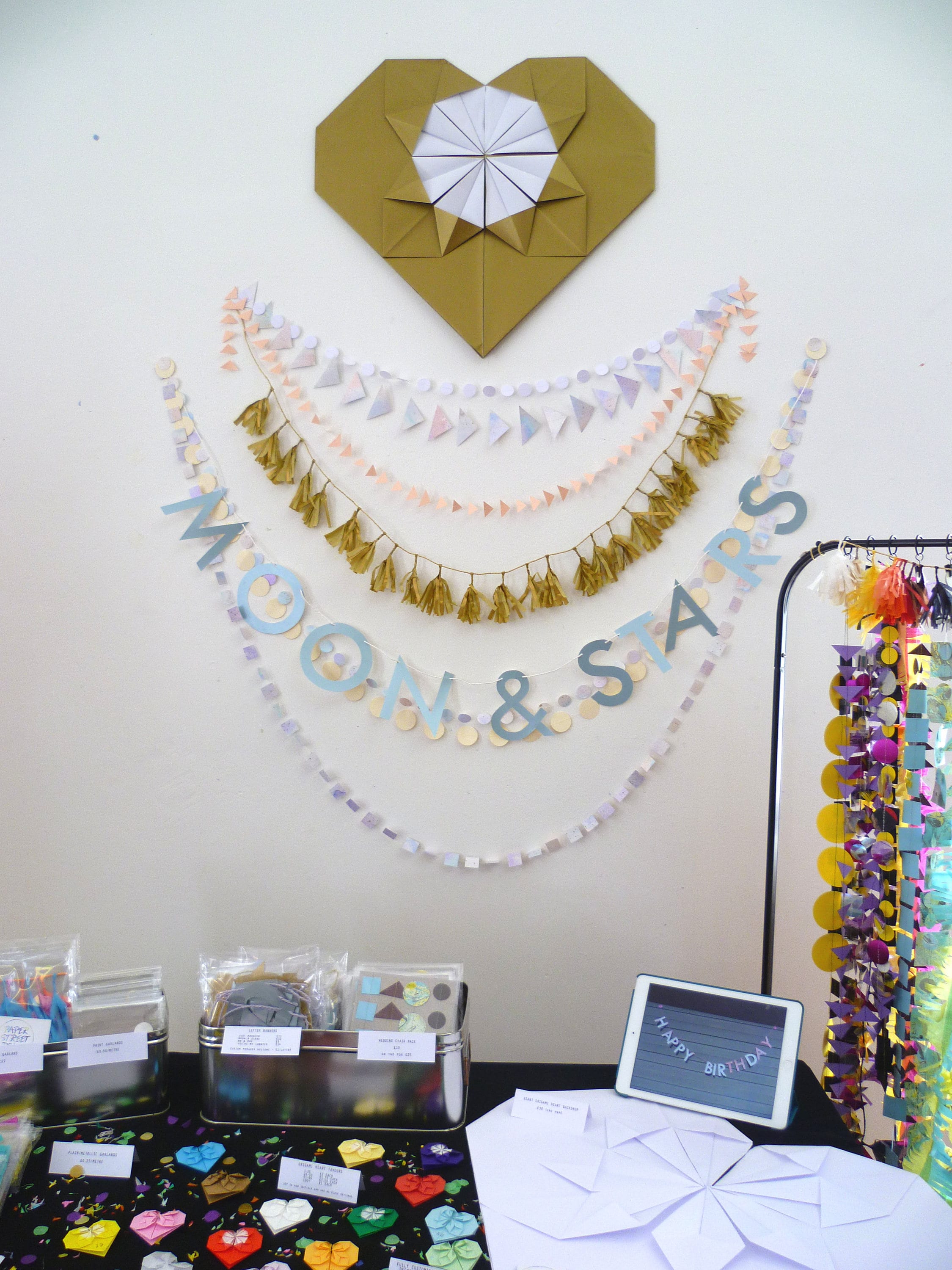 Wedding wall display at a wedding fair on Buccleuch St. in Edinburgh, Scotland. A giant gold origami heart; below several strand of pastel paper garland; tiny gold tissue tassel garland; more strands of paper garland and a banner that reads "MOON & STARS"