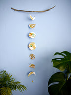 Moon phases stitched garland painted with green and ochre inks hanging from a piece of driftwood displayed on a blue wall with plants at either side