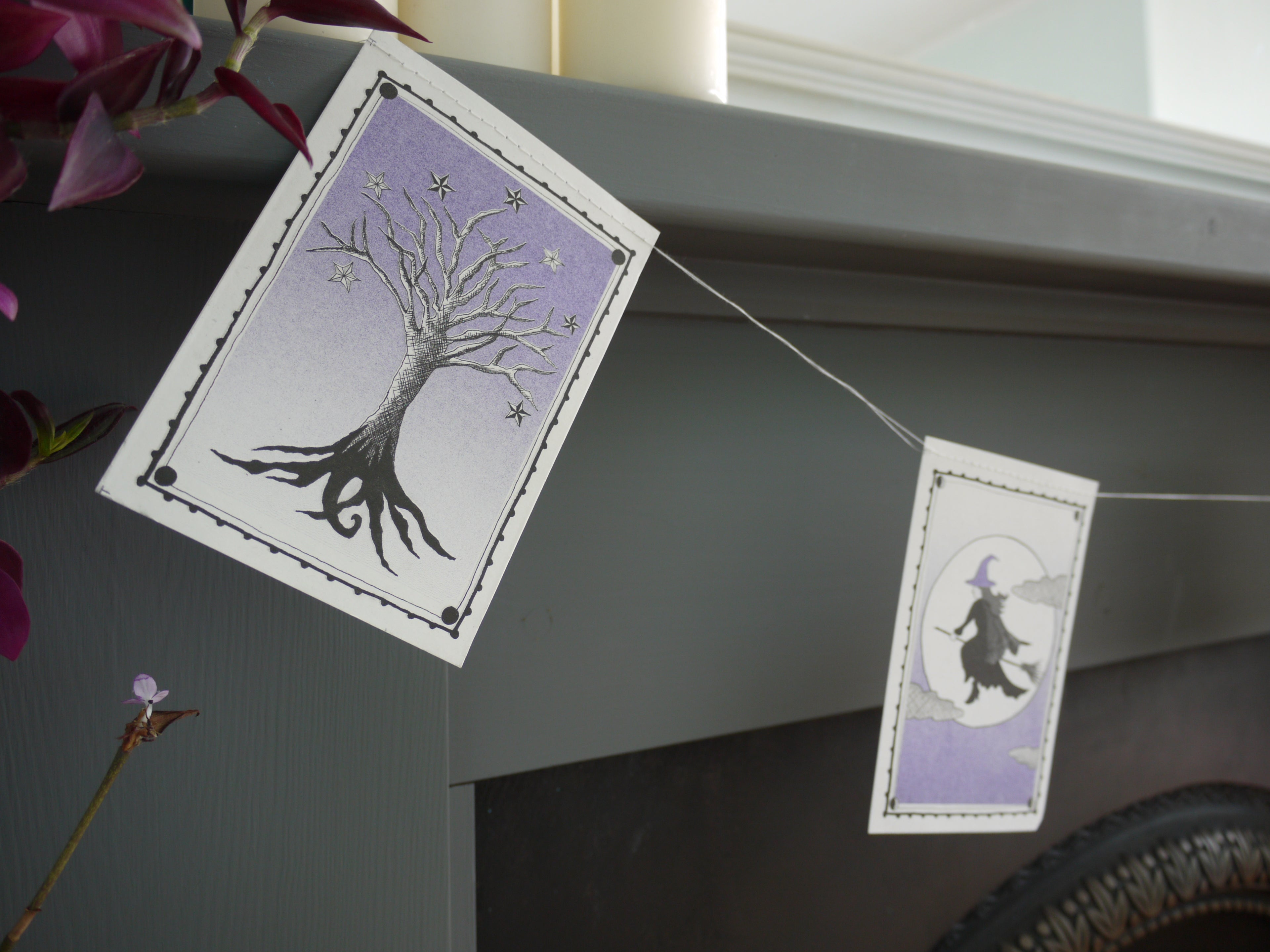 Close up of an illustrated tree and a witch riso printed stitched Halloween paper garland in black white and purple hanging above a grey and black fireplace