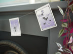 Close up of a spider and bats illustrated riso printed stitched Halloween paper garland in black white and purple hanging above a grey and black fireplace with a pink plant ant the right side