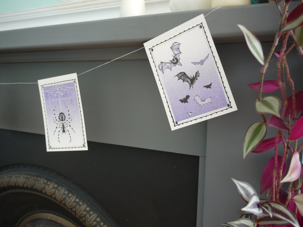 Close up of a spider and bats illustrated riso printed stitched Halloween paper garland in black white and purple hanging above a grey and black fireplace with a pink plant ant the right side