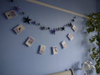 Illustrated riso printed stitched Halloween paper garland in black white and purple hanging on a blue wall with purple, black and grey star garland hanging above with a white lamp and green plant at the right side