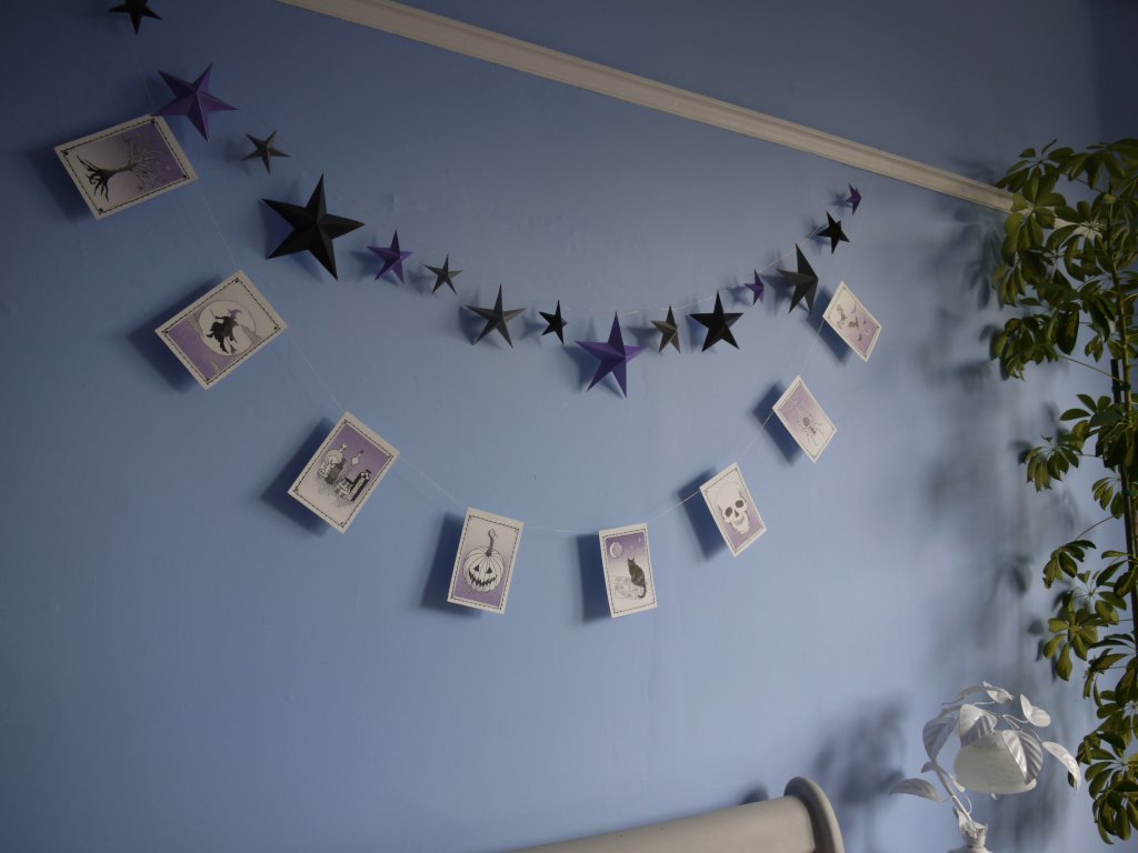 Illustrated riso printed stitched Halloween paper garland in black white and purple hanging on a blue wall with purple, black and grey star garland hanging above with a white lamp and green plant at the right side