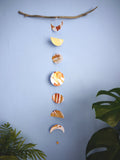 Moon phases stitched garland painted with golden tans and brown ink stitched to create a wall hanging displayed on a blue background from a piece of driftwood with green plants on either side