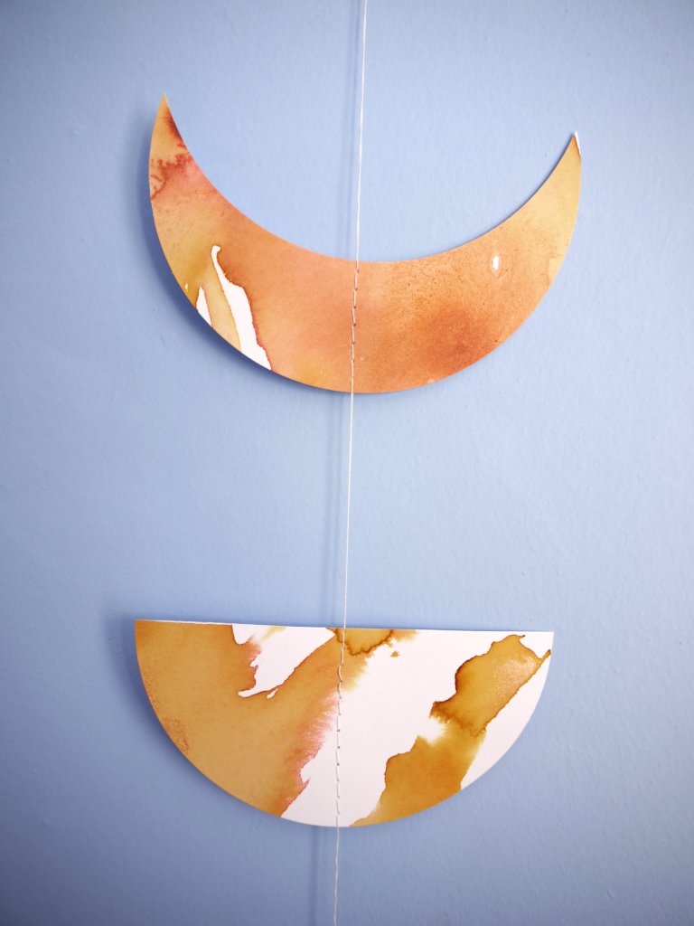 Close up of moon phases stitched garland painted with golden tan and brown inks displayed as a wall hanging on a blue background 