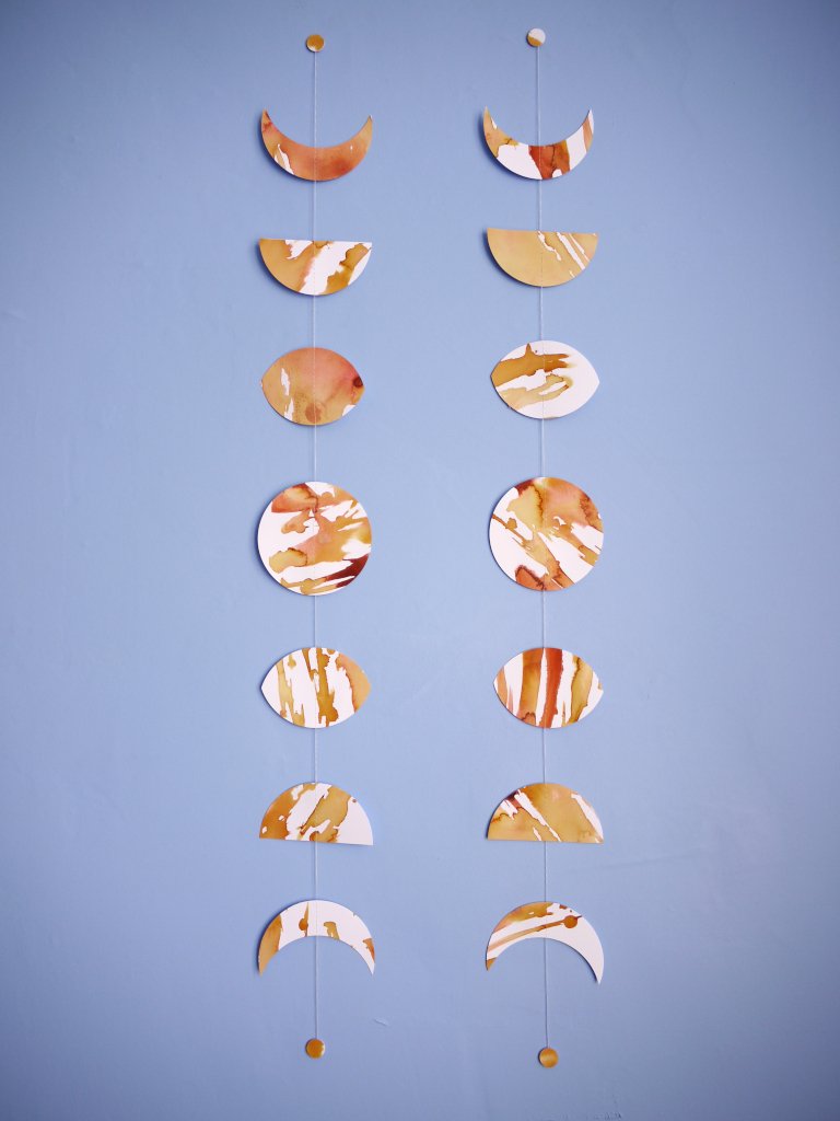 Two moon phases stitched garlands painted slightly differently with golden tan and brown inks displayed as a wall hanging on a blue background 