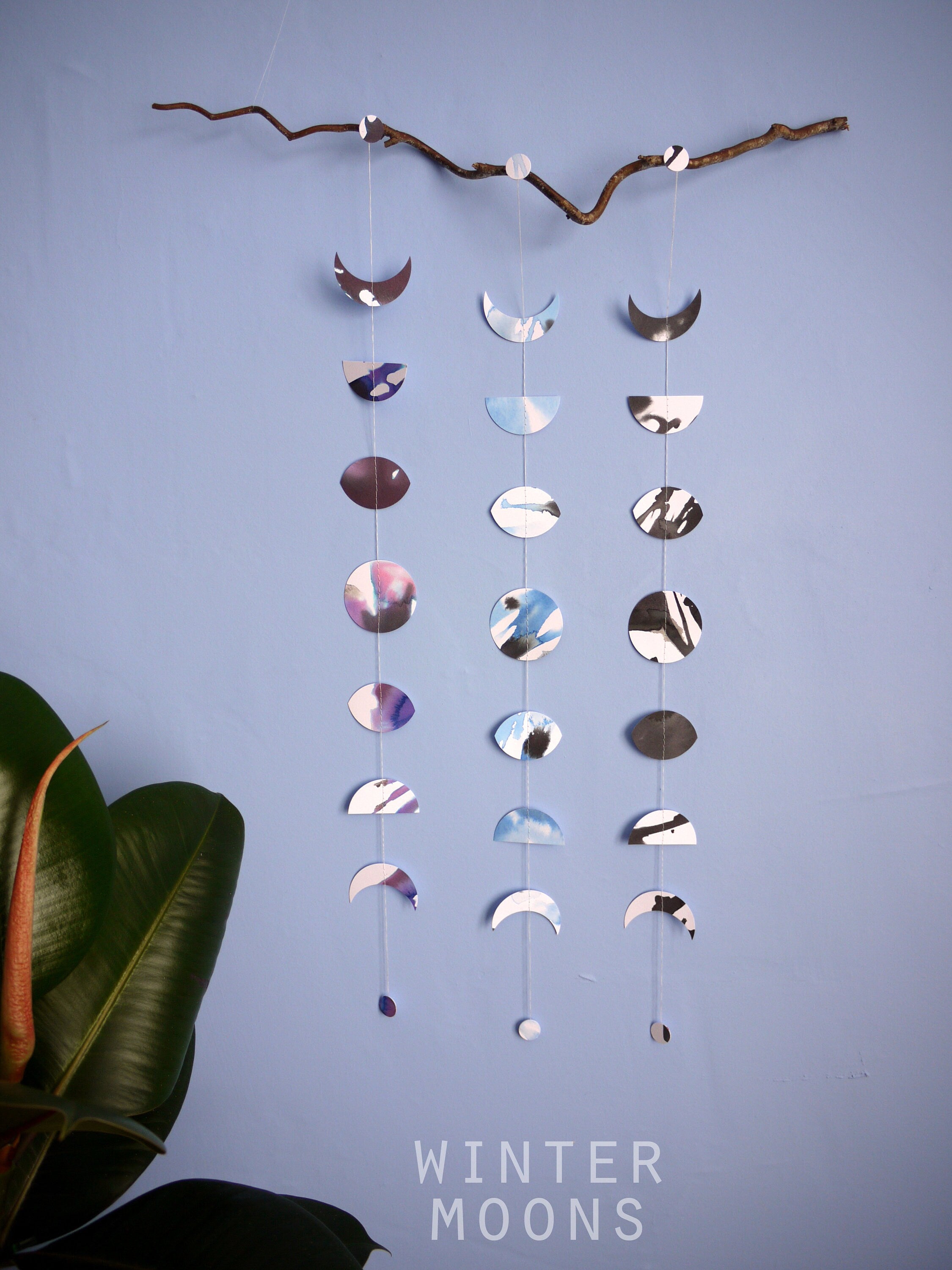 Wall hanging of 3 mini stitched paper garlands depicting the phases of the moon each painted with different colours of ink to show different months of the year hanging vertically from a piece of driftwood on a blue wall with a plant at the left side and text at the bottom saying winter moons