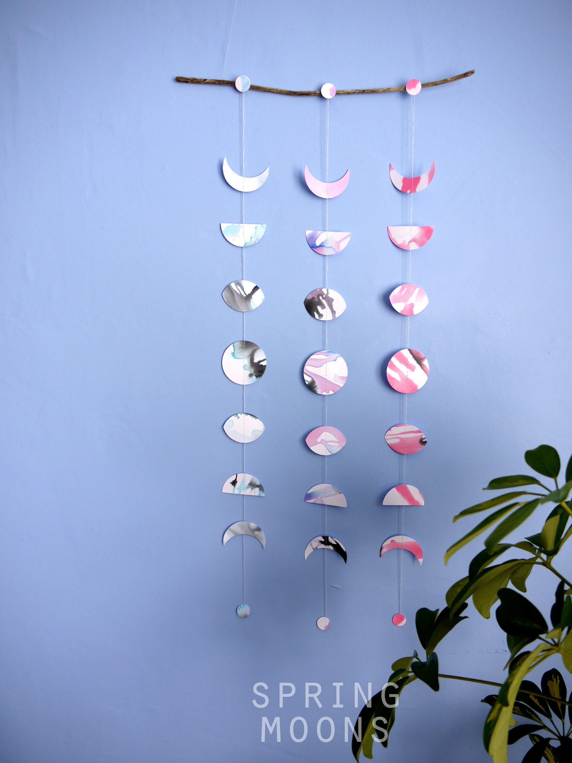 Wall hanging of 3 mini stitched paper garlands depicting the phases of the moon each painted with different colours of ink to show different months of the year hanging vertically from a piece of driftwood on a blue wall with a plant at the right side and text at the bottom saying spring moons