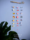 Wall hanging of 3 mini stitched paper garlands depicting the phases of the moon each painted with different colours of ink to show different months of the year hanging vertically from a piece of driftwood on a blue wall with a plant at the left side and text at the bottom saying summer moons