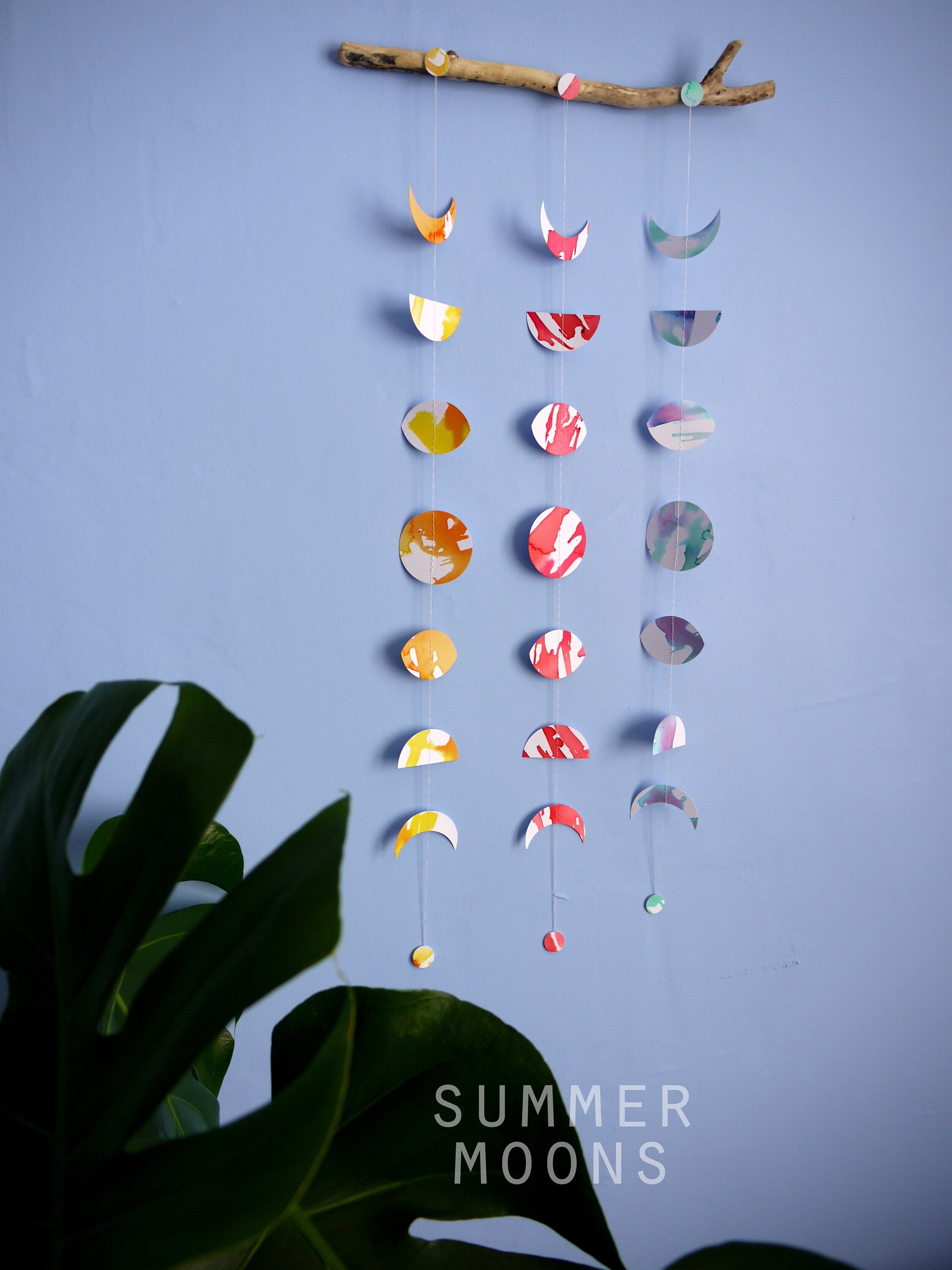 Wall hanging of 3 mini stitched paper garlands depicting the phases of the moon each painted with different colours of ink to show different months of the year hanging vertically from a piece of driftwood on a blue wall with a plant at the left side and text at the bottom saying summer moons