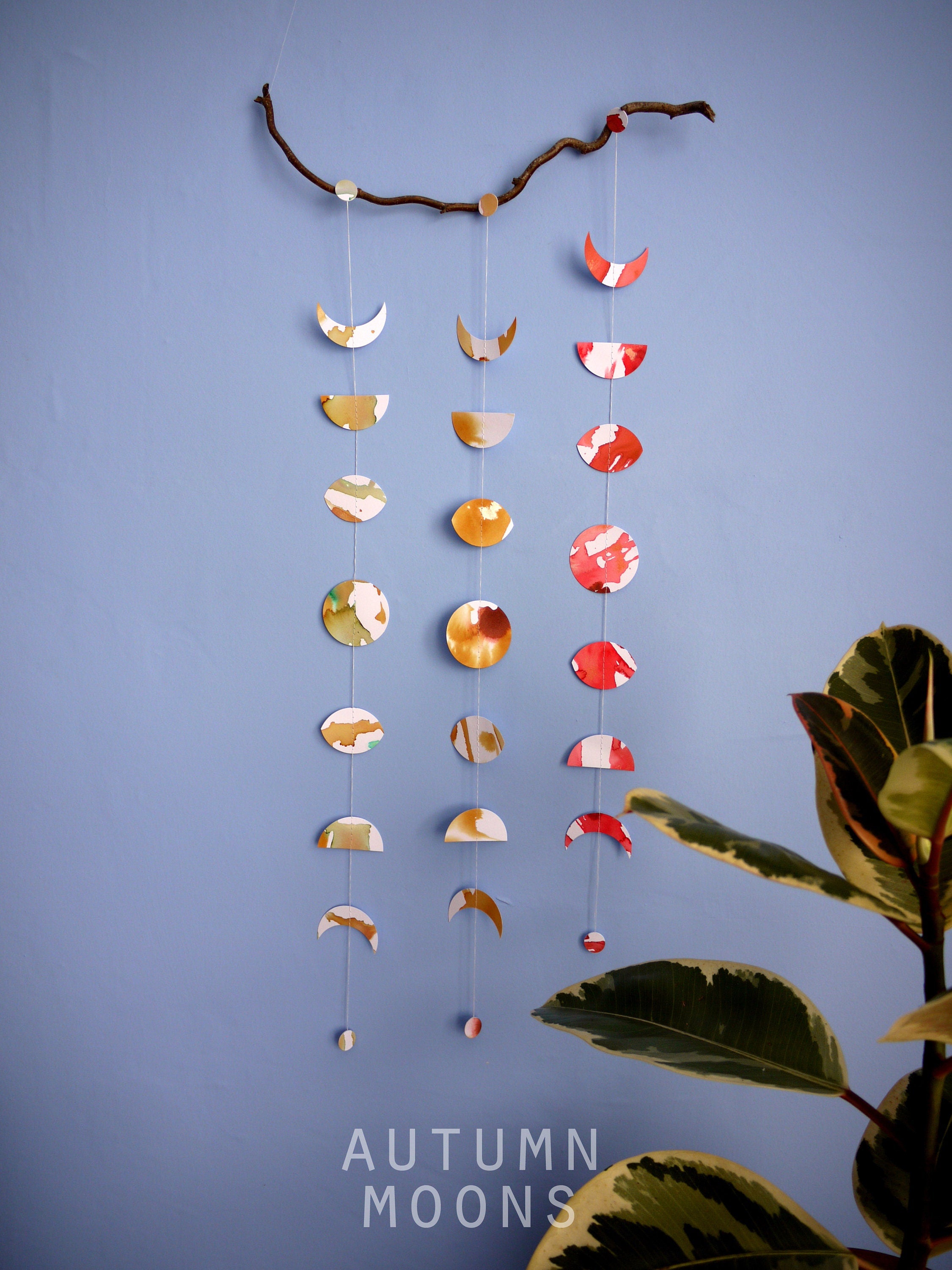 Wall hanging of 3 mini stitched paper garlands depicting the phases of the moon each painted with different colours of ink to show different months of the year hanging vertically from a piece of driftwood on a blue wall with a plant at the right side and text at the bottom saying autumn moons