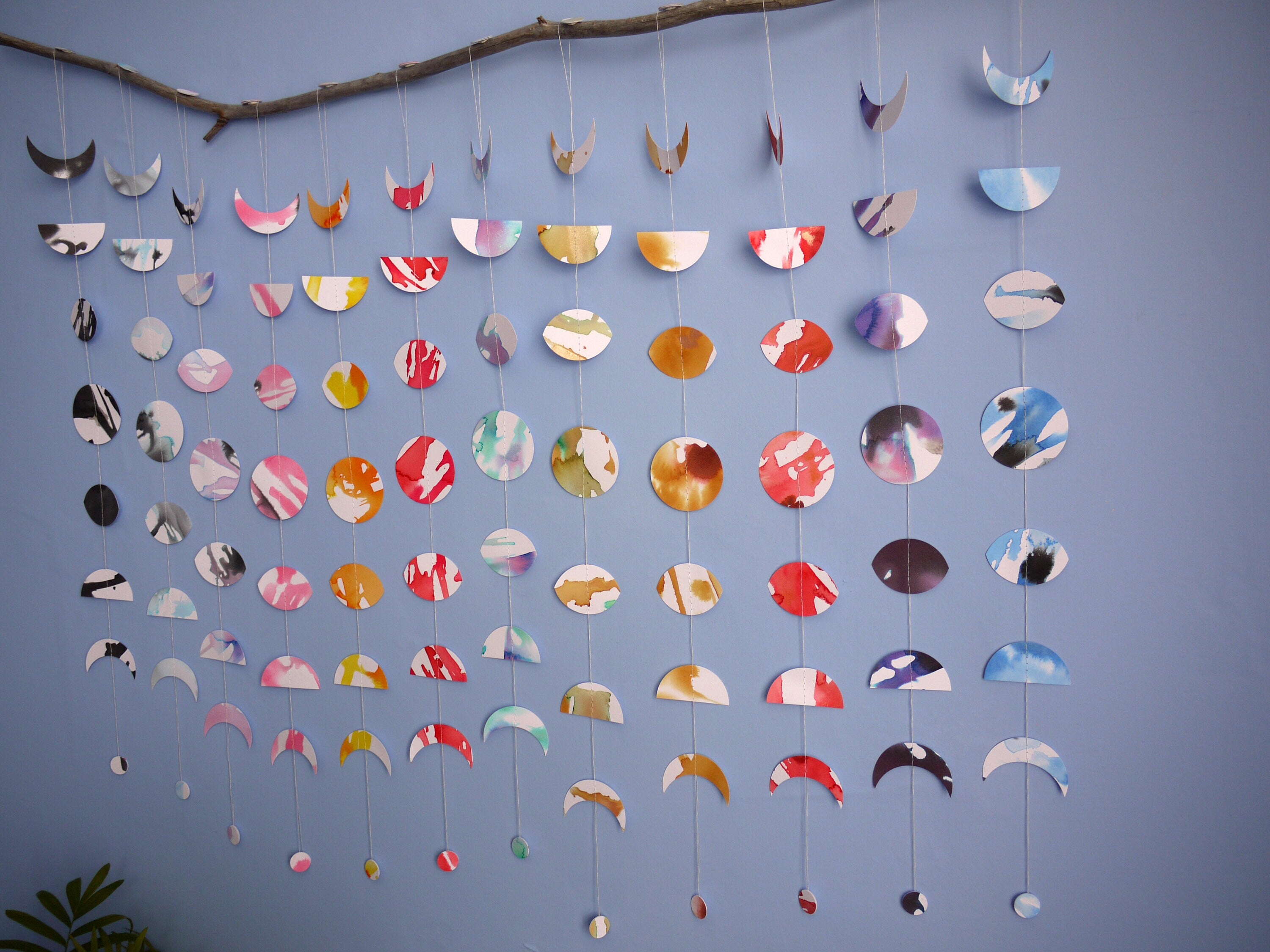 Wall hanging of 12 mini stitched paper garlands depicting the phases of the moon each painted with different colours of ink to show each month of the year hanging vertically from a piece of driftwood on a blue wall