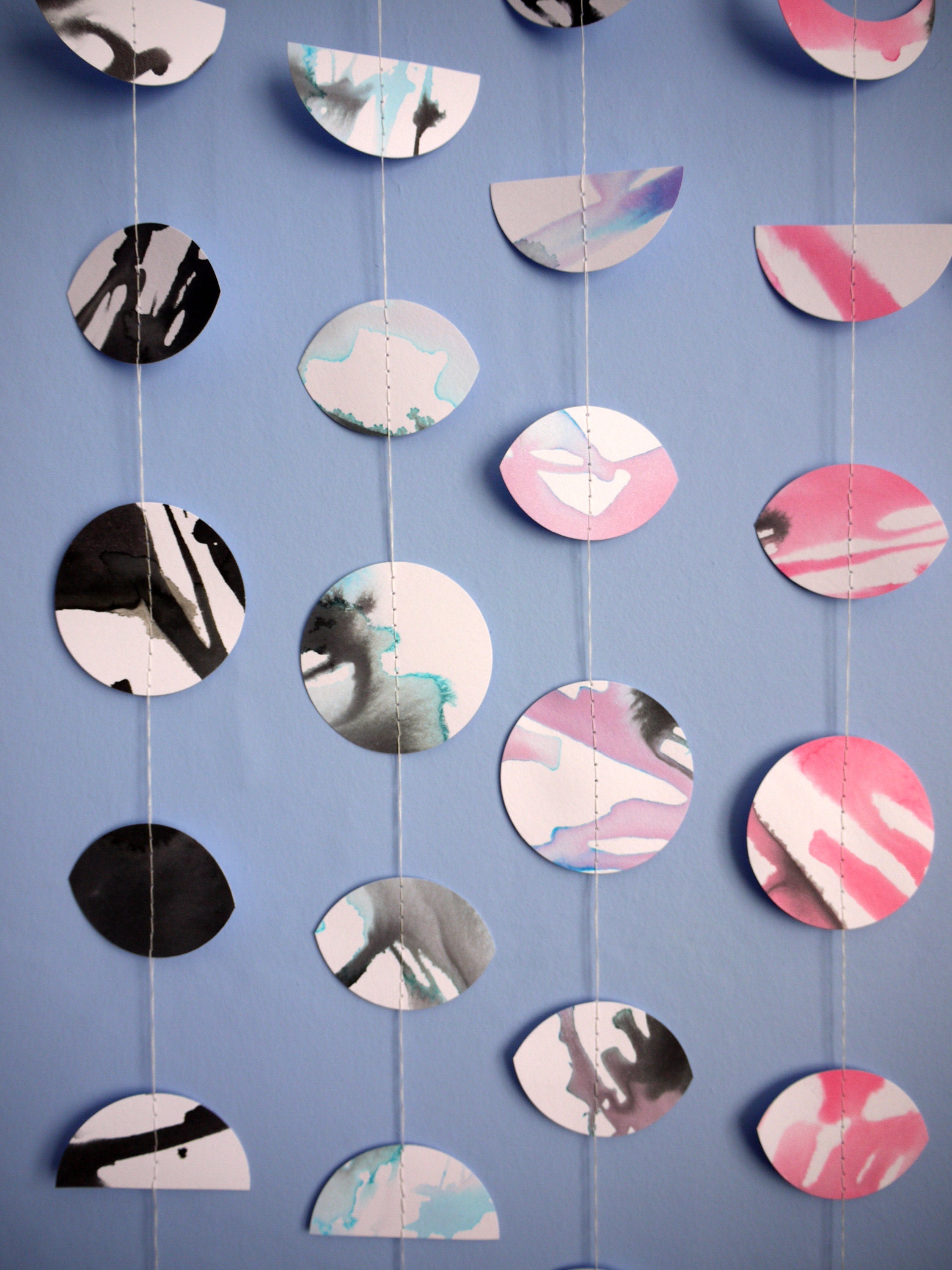 Wall hanging of 4 mini stitched paper garlands depicting the phases of the moon each painted with different colours of ink to hanging vertically on a blue wall