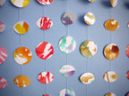 Wall hanging of mini stitched paper garlands depicting the phases of the moon each painted with different colours of ink to show different months of the year hanging vertically on a blue wall
