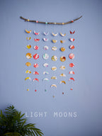 Wall hanging of 6 mini stitched paper garlands depicting the phases of the moon each painted with different colours of ink to show different months of the year hanging vertically from a piece of driftwood on a blue wall with a plant at the left side and text at the bottom saying light moons