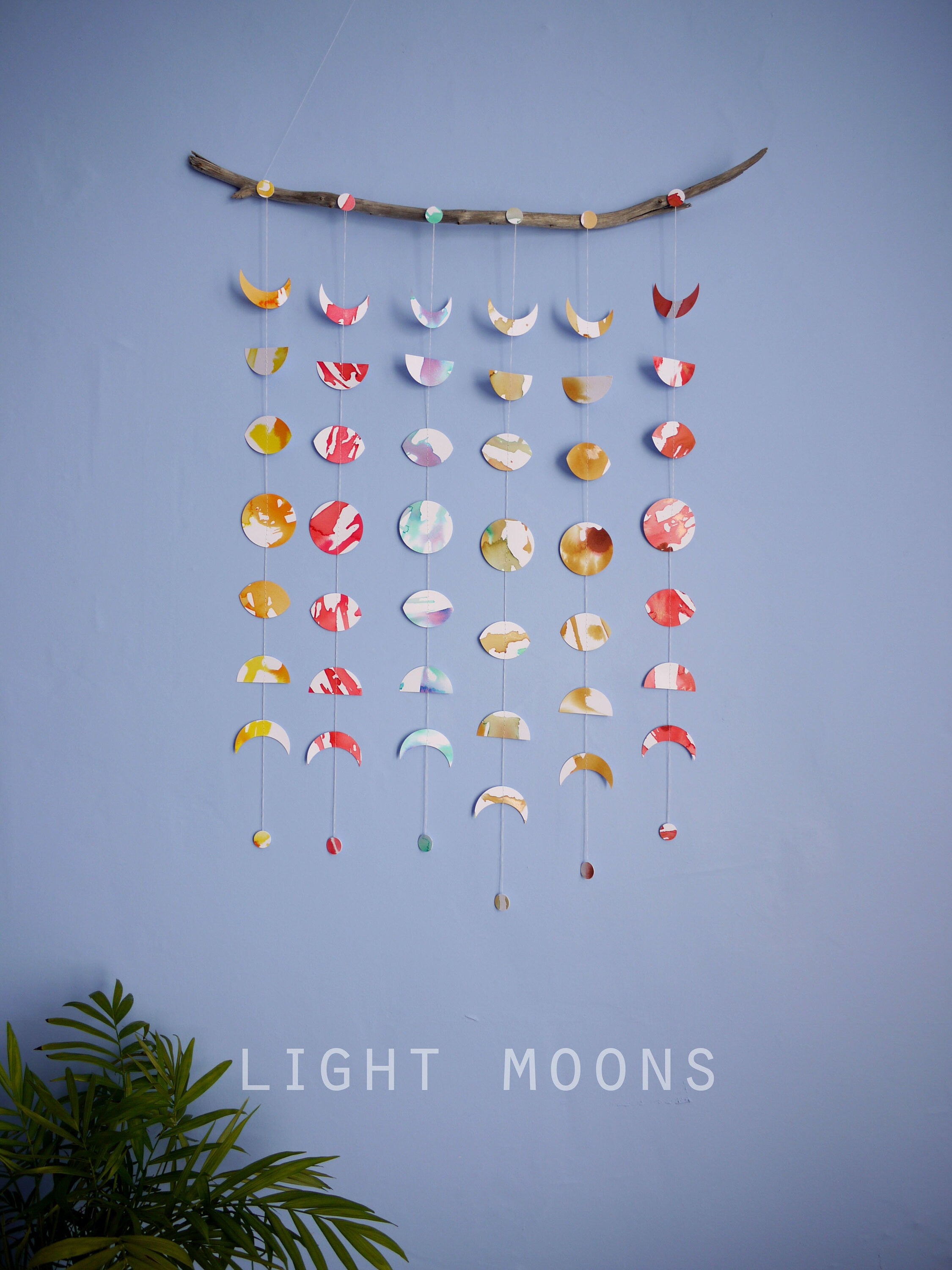 Wall hanging of 6 mini stitched paper garlands depicting the phases of the moon each painted with different colours of ink to show different months of the year hanging vertically from a piece of driftwood on a blue wall with a plant at the left side and text at the bottom saying light moons