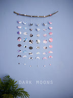 Wall hanging of 6 mini stitched paper garlands depicting the phases of the moon each painted with different colours of ink to show different months of the year hanging vertically from a piece of driftwood on a blue wall with a plant at the left side and text at the bottom saying dark moons