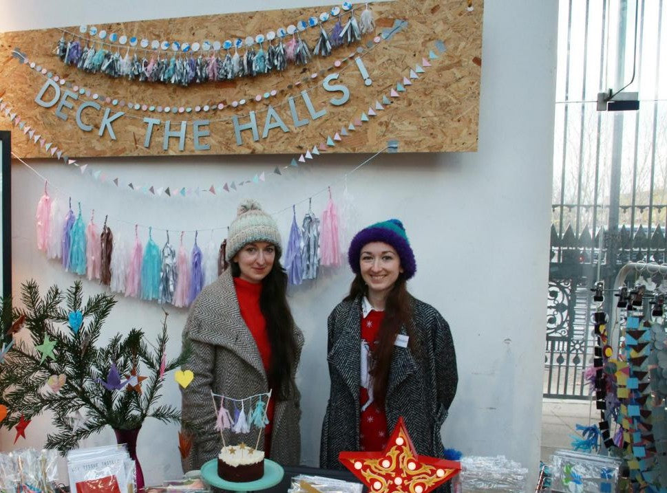 Two women standing in front of a Christmas-themed display with decorations and a sign that says 'Deck the Halls!'.