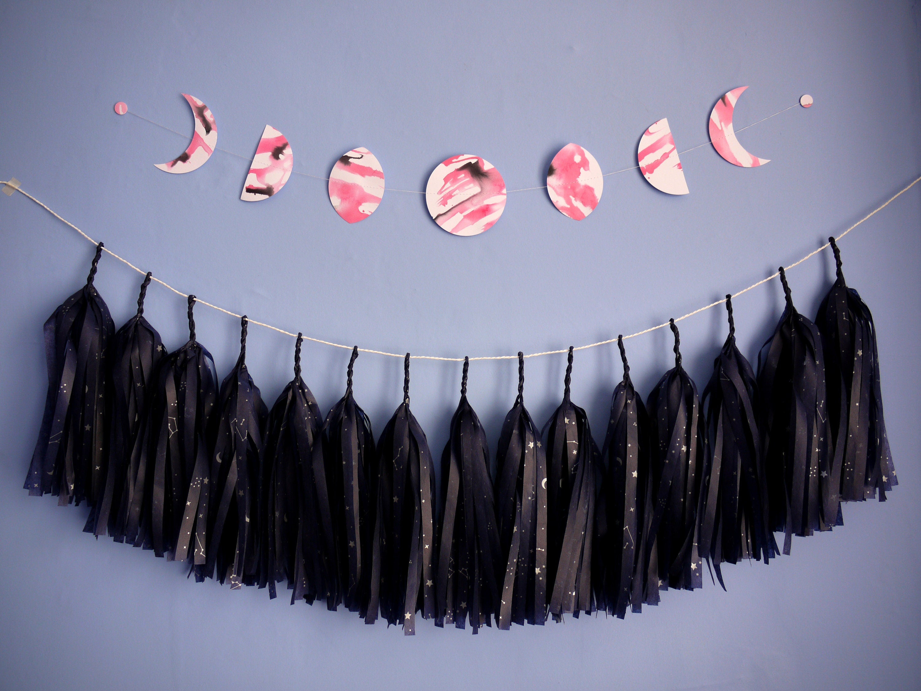 Moon phase shapes painted with pink and black ink stitched to create a wall hanging displayed on a blue background with navy blue constellation printed tissue paper tassel garland hanging underneath 