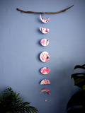 Vertical moon phase shapes painted with pink and black ink stitched to create a wall hanging displayed hanging from a piece of driftwood on a blue background with plants on either side