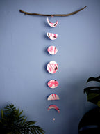 Vertical moon phase shapes painted with pink and black ink stitched to create a wall hanging displayed hanging from a piece of driftwood on a blue background with plants on either side