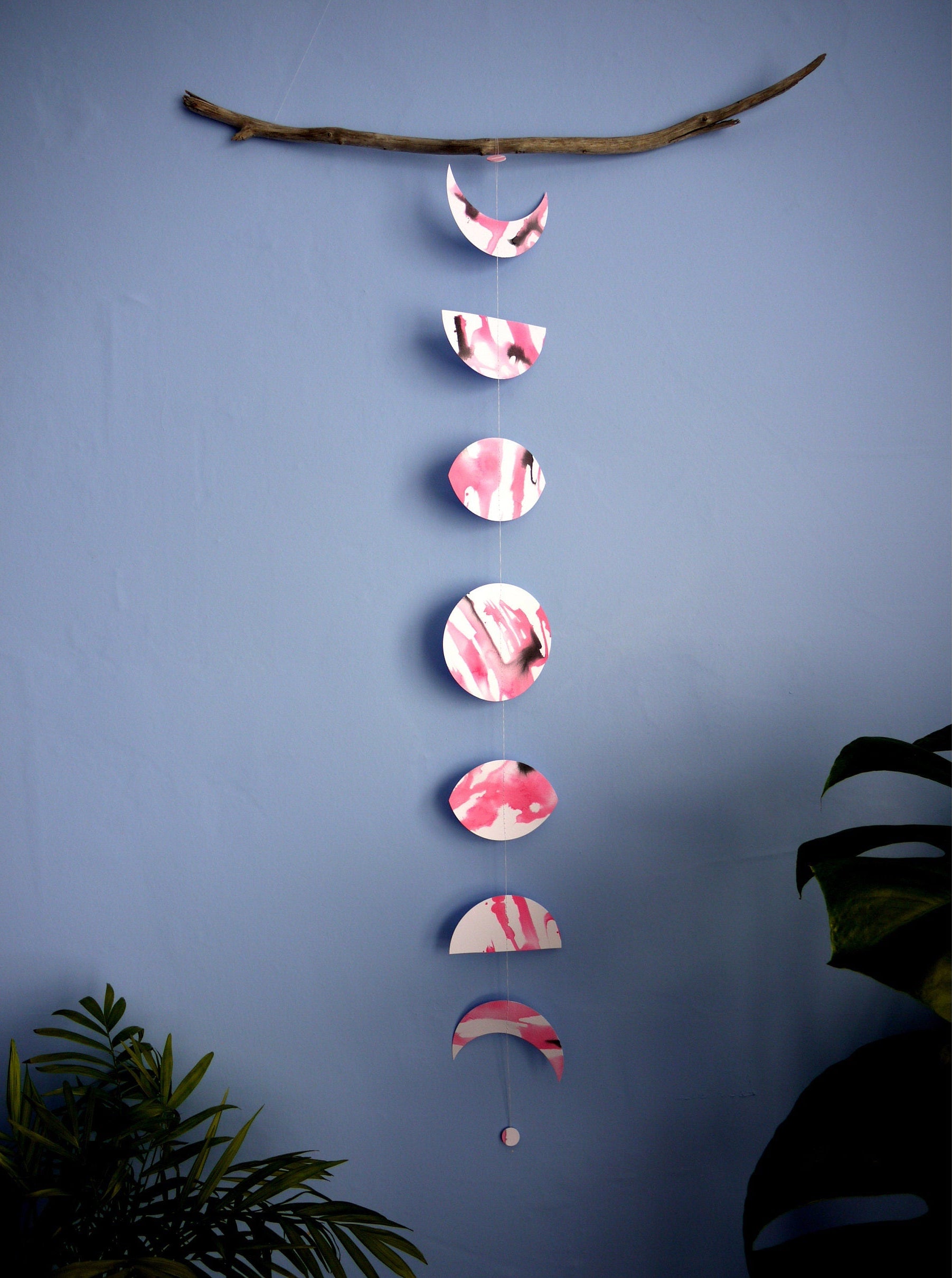 Vertical moon phase shapes painted with pink and black ink stitched to create a wall hanging displayed hanging from a piece of driftwood on a blue background with plants on either side