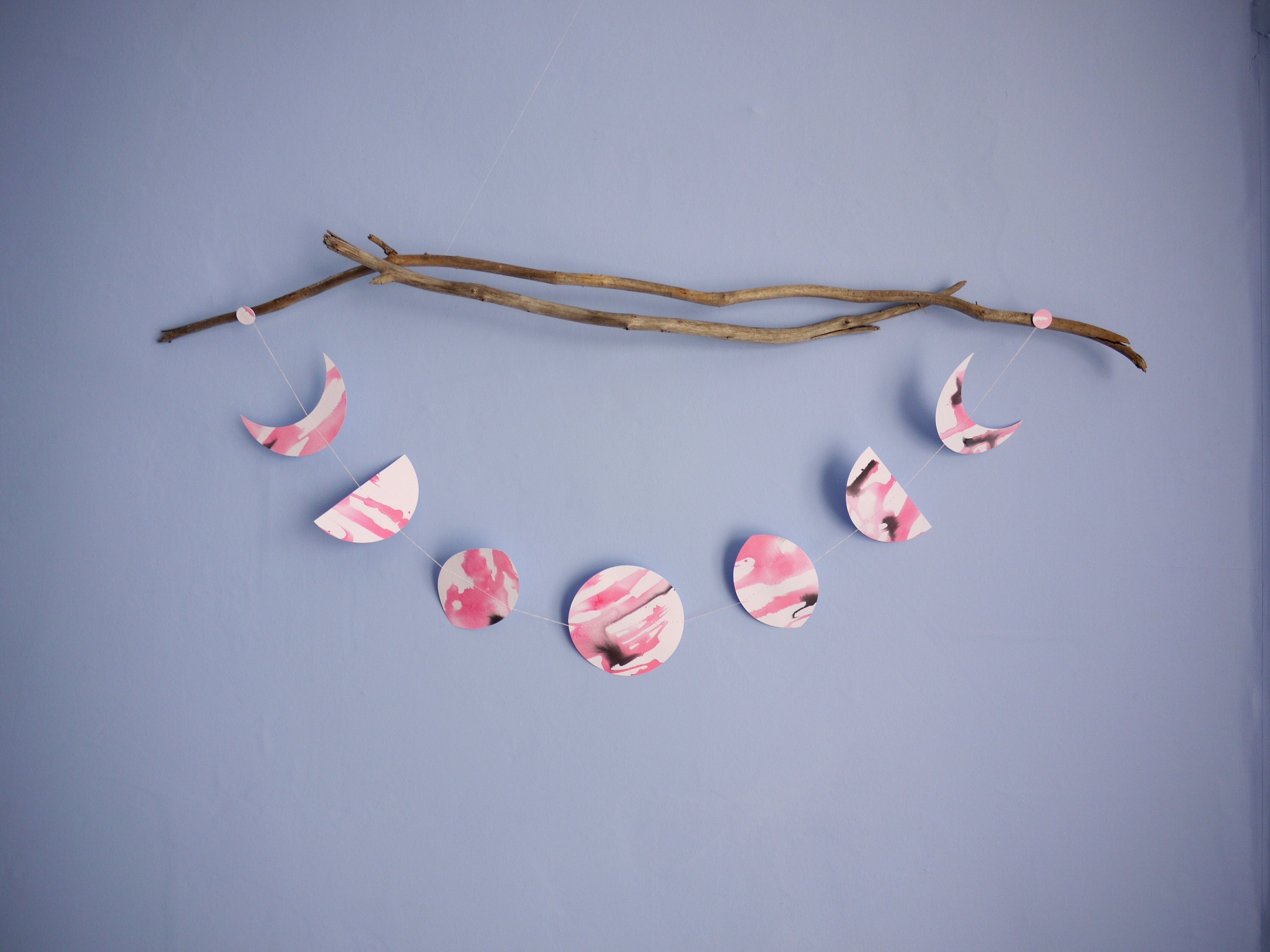 Horizontal moon phase stitched paper garland painted with pink and black ink displayed from driftwood on a blue background 